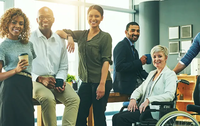 Group of six diverse professionals smiling in an office environment, one of whom is using a wheelchair.